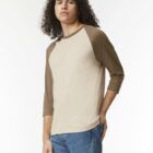 Man with curly hair in a tan and brown raglan shirt and blue jeans standing against white background.