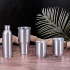 Four stainless steel drinkware items on a wooden surface with a plant in the background.