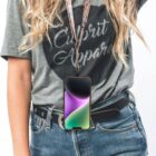 Woman with wavy blonde hair wearing a gray t-shirt that says Calipri Apparel and black jeans. She has a smartphone with a colorful screen hanging from a lanyard around her neck. Hands are partially visible, fingers in jeans pocket. White background.