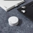 A white sound machine on a desk beside earbuds, a laptop, and a notebook.