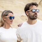 Man and woman wearing sunglasses, standing in front of a sandy background.