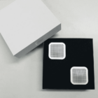 Two square, perforated white devices in an open black box next to a white lid.