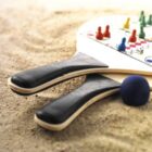Two beach paddles with black handles and a colorful game board design rest on sand. A blue ball lies next to them.