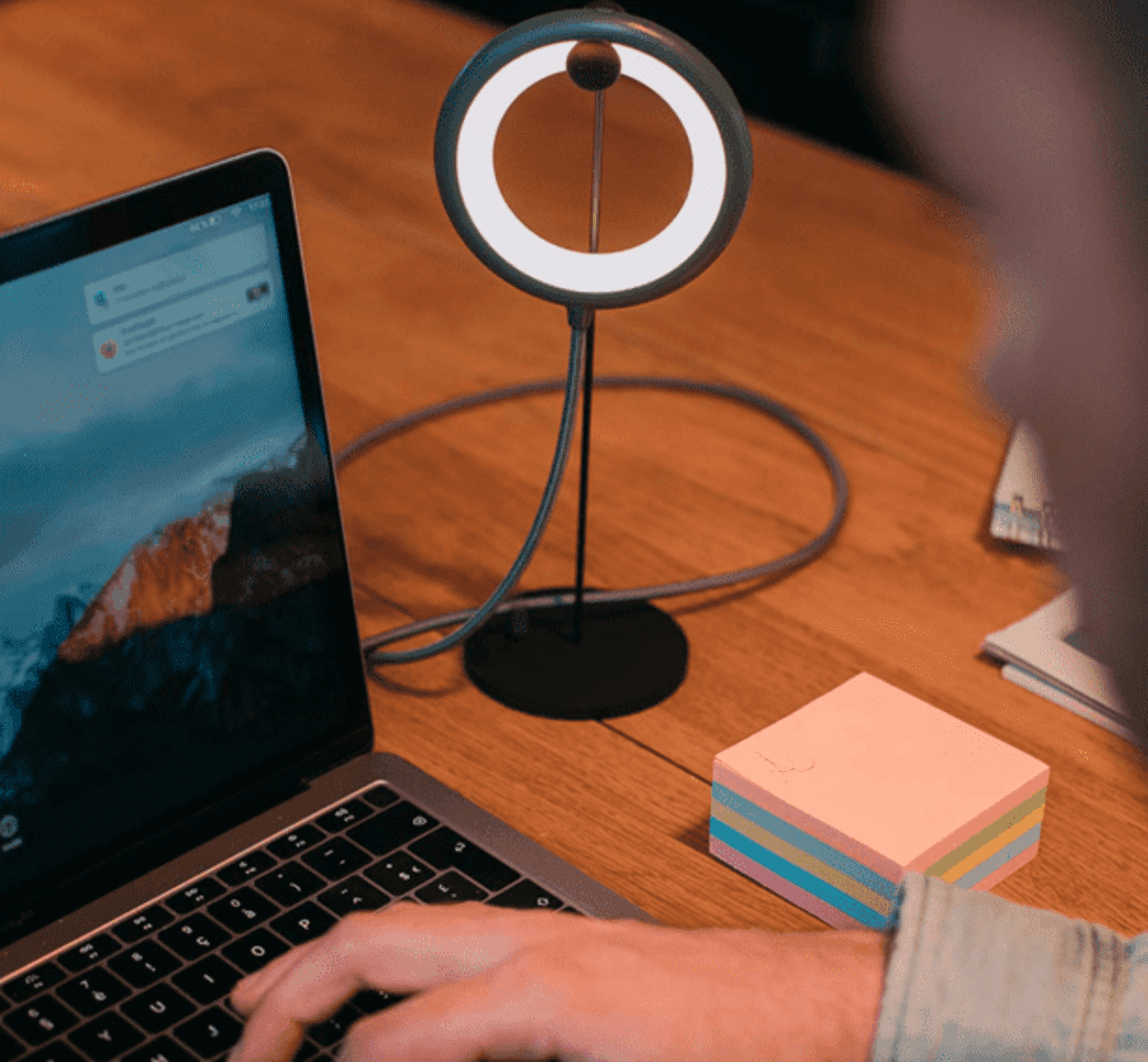 Person typing on laptop near a ring light and sticky notes on a wooden desk.