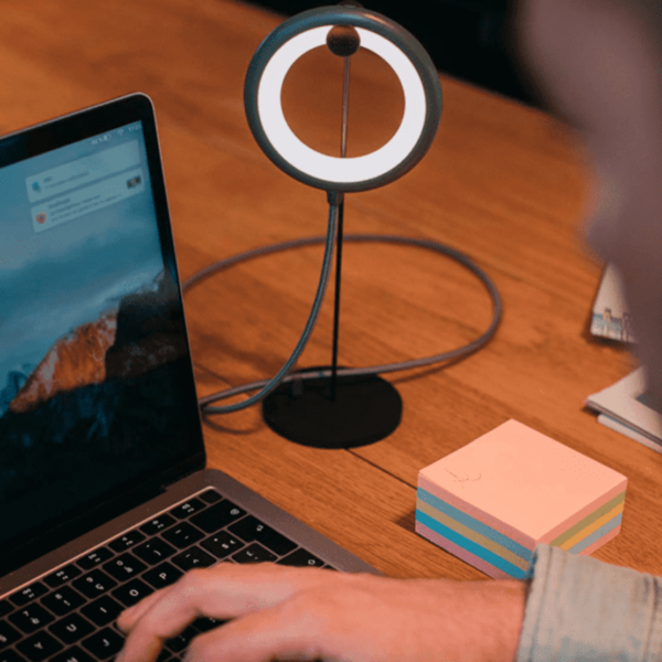 Person using a laptop at a desk with a small ring lamp and sticky notes nearby.