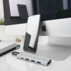 A smartphone leans against a computer, with USB hubs and a plant nearby.