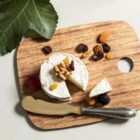 Round brie cheese topped with walnuts on a wooden board, surrounded by almonds, raisins, cranberries, raspberries, and blackberries. A slice is cut out, and a silver knife with a gold blade rests nearby. Large green leaf partially visible in the corner.