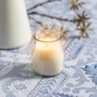 A lit white candle in a small glass jar on a blue-and-white patterned surface, with an out-of-focus plant in the background.