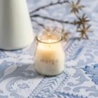 A lit candle in a small glass jar with morga written on it sits on a patterned blue and white surface. Out-of-focus branches with star-shaped decorations are in the background, alongside a white vase.