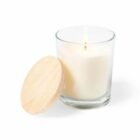 A lit white candle in a clear glass jar is shown with a wooden lid placed beside it on a white background.