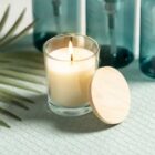 A lit white candle in a clear glass jar sits on a textured surface with a wooden lid nearby. Blue bottles are in the background, and a palm leaf is placed to the side.
