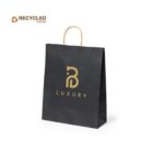 Black paper bag with gold handle, featuring a stylized B logo and the word LUXURY in gold. RECYCLED PAPER is written in the top left corner with a recycling symbol.