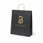 A black shopping bag with gold handles and a gold logo featuring the letter B and the word LUXURY in capital letters on the front. The bag is standing upright against a plain white background.