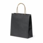 A simple black paper shopping bag with brown twisted handles, standing upright on a plain white background. The bag is slightly open at the top, showcasing its minimalistic design.