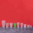 Eight cups of varying sizes, one green among clear cups, lined up on a gray surface, red background.