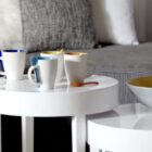 Three white mugs with colored spoons on a round white table near a gray sofa and a bowl.