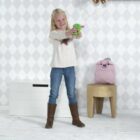 A young girl with long blonde hair is joyfully blowing bubbles using a green bubble blower. She is wearing a white sweater, blue jeans, and brown boots. Behind her are toys on a shelf and a pink backpack on a stool. The room has a white patterned wall.
