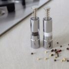 Two metallic pepper mills on a countertop with scattered peppercorns nearby.