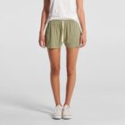 Person in a white shirt and green shorts standing, wearing white sneakers, against a plain backdrop.