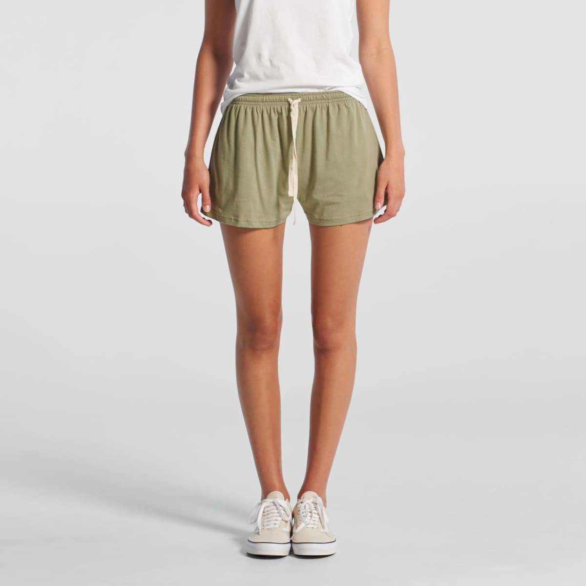 Person in a white shirt and green shorts standing, wearing white sneakers, against a plain backdrop.