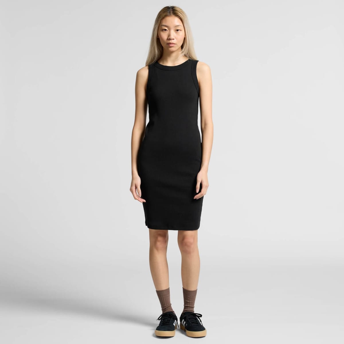 Woman in a sleeveless black dress and sneakers stands against a plain light background.