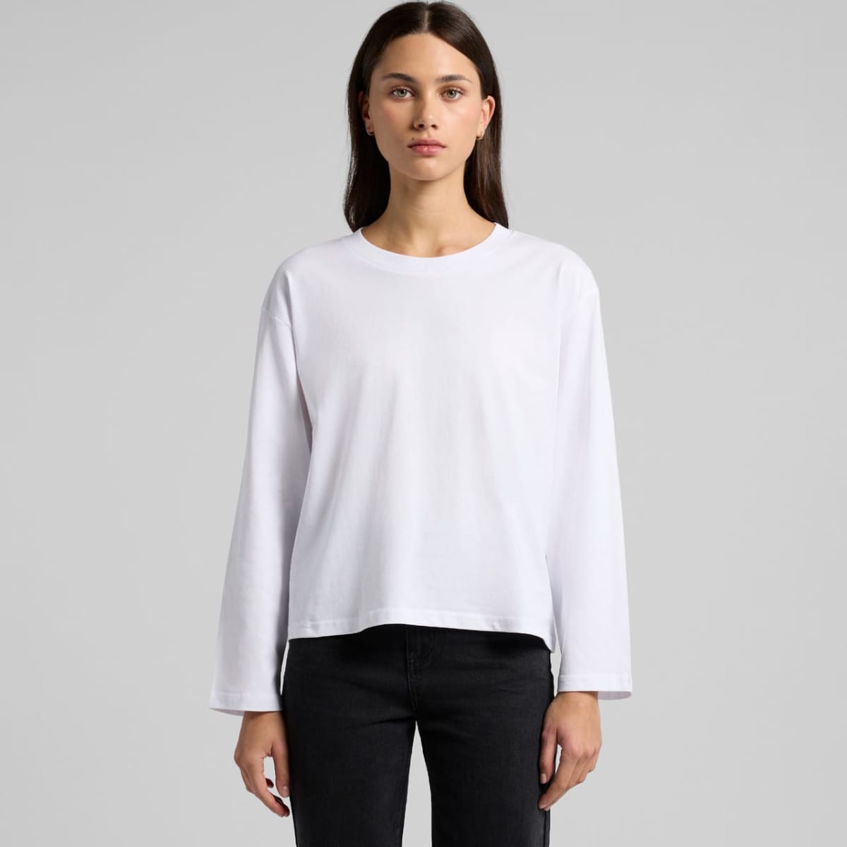 Woman wearing a plain white long-sleeve shirt and black pants, standing against a gray background.