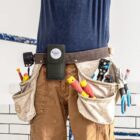 Person wearing a tool belt with various tools in front of a white wall.