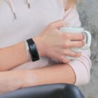 Person in a pink hoodie holding a white mug, wearing a black fitness tracker and silver bracelet.
