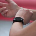 Person wearing a black smartwatch, raising their other hand against a blurred red and white background.