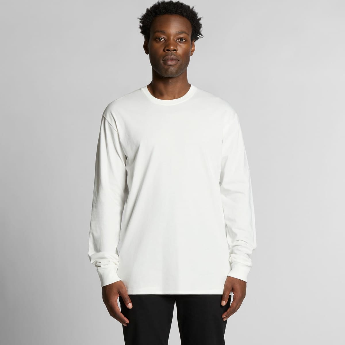 Person in a plain white long-sleeve shirt and black pants stands against a gray background.