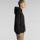 Person in black hoodie and khaki pants standing in profile against a gray background.