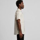 Person in a beige short-sleeve shirt and black pants, side view, against a gray background.