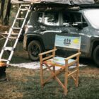 Camping scene with a dark green SUV parked near trees at twilight. A rooftop tent is set up on the vehicle with a ladder. A folding chair with Summer Breeze and a sun design is placed on the grass, near a small campfire.