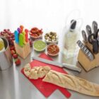 Kitchen setup with knives, wine, sliced baguette, and various small dishes on a counter.