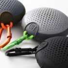 Three round portable speakers with carabiners in green, orange, and black on a white surface.
