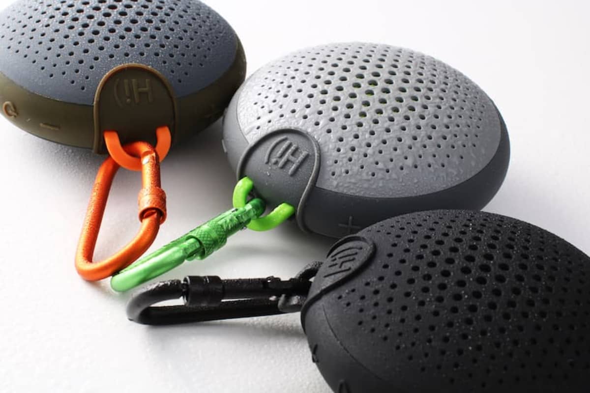 Three round portable speakers with carabiners in green, orange, and black on a white surface.
