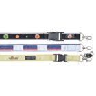 Three lanyards with various designs. The top one is black with colorful circular symbols, the middle one is white with blue text, and the bottom one is light yellow with a logo. Each has a metal clip and a plastic buckle.