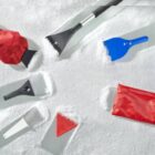 Various ice scrapers and mitts on snow, including red, blue, white, and black tools.