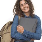 Smiling person with curly hair, wearing a denim shirt, holds a folder and has a backpack on one shoulder, standing against a white background.
