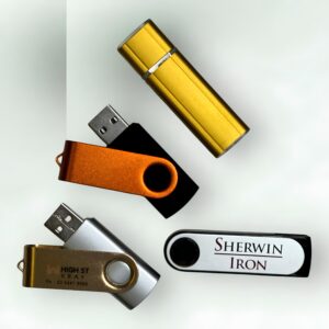 Four promotional USBs are displayed against a light background. One is gold and cylinder-shaped, while three feature swivel designs: an orange and black, a sleek silver, and a classic white bearing the Sherwin Iron logo.