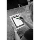 A person holding a white tablet with both hands, using a green stylus to interact with the screen. The background features a marble or granite table and empty chairs. The image is in black and white, except for the green stylus.