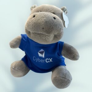 A plush hippopotamus toy, a charming example of promotional products, dons a blue t-shirt featuring a geometric logo and the text CyberCX. With its friendly expression set against a softly blurred background, it captures both warmth and brand spirit.