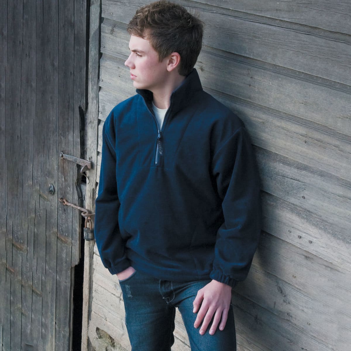 Young man in a navy pullover leans against a wooden wall, looking to the left.