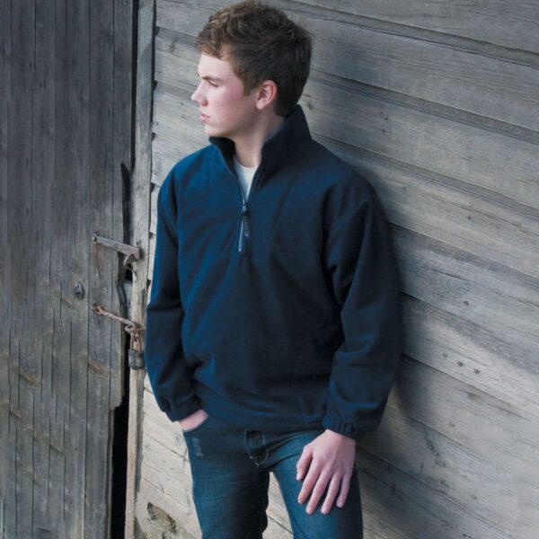 A person in a navy pullover leans against a wooden wall, looking to the left.