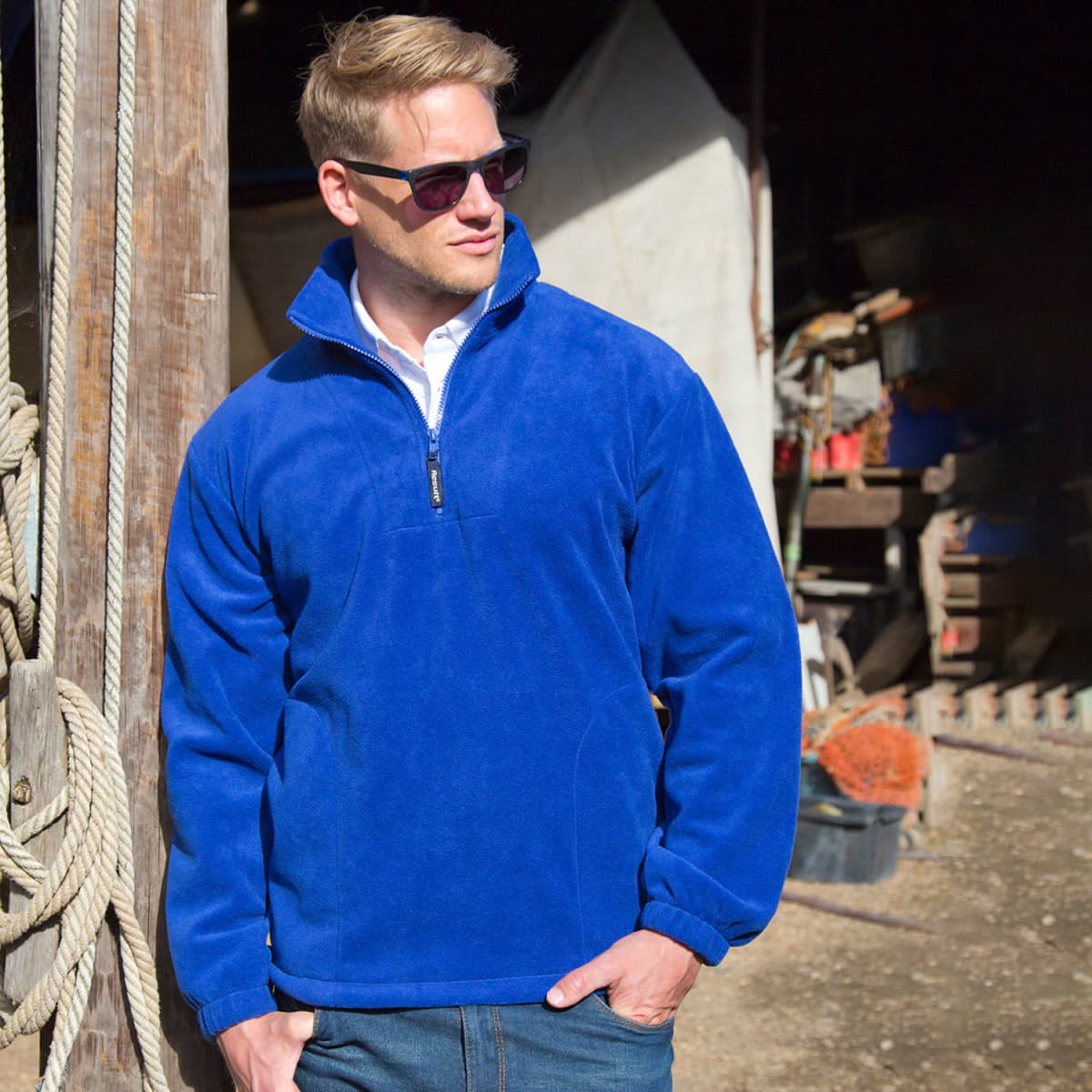 Man in blue fleece jacket and sunglasses standing outdoors near ropes and wooden structure.