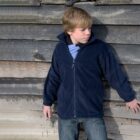 Boy in a navy jacket stands against a weathered wooden wall, looking to the side.