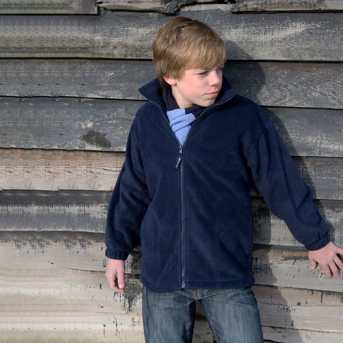 Boy in a navy jacket stands against a weathered wooden wall, looking to the side.