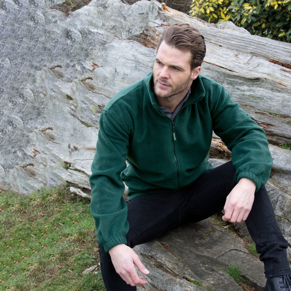 Man in a green jacket sits on a large rock outdoors, looking thoughtfully to the side.