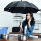 Woman indoors using phone, holding an umbrella under leaking ceiling.