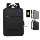 Three sleek backpacks in black and gray with a built-in USB charging port are displayed. One backpack shows a phone connected, indicating charging functionality. The design features a top handle and multiple zippered compartments.